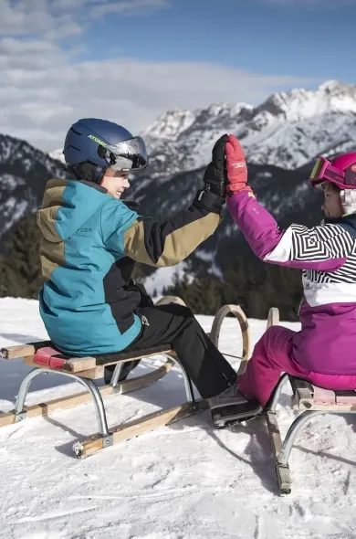 Zwei Kinder auf Holzrodel sitzend von hinten